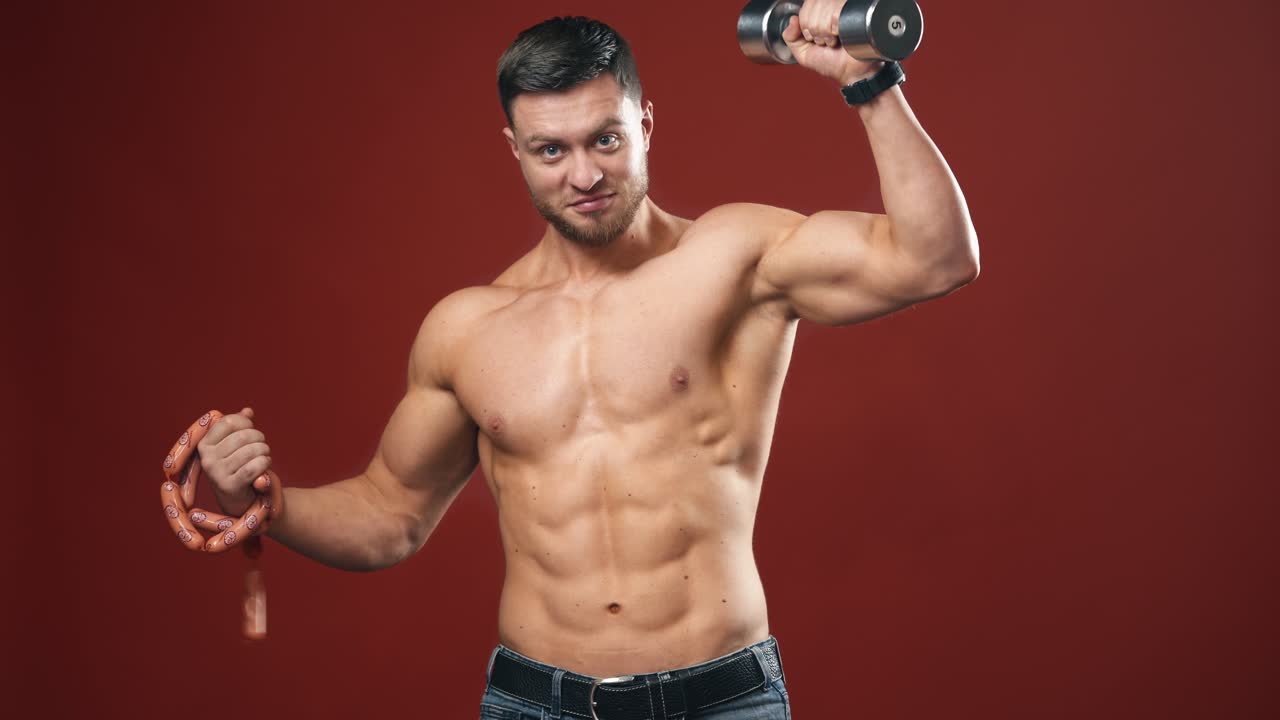 Athlete male with dumbbell and sausage. Young sportsman chooses between heavy dumbbell and sausage. Shirtless bodybuilder on the red sturdio background.
