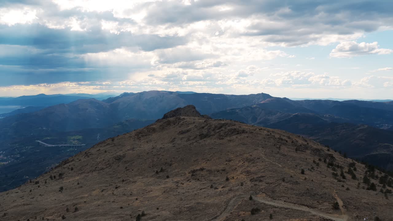 Aerial footage from Mount Praglia showing rugged peaks under dramatic cloud cover, with sun rays piercing through the sky and a distant glimpse of the Ligurian Sea and mountain valleys
