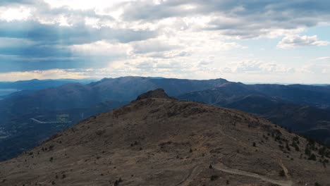 aerial footage from mount praglia showing rugged peaks under dramatic cloud cover, with sun rays piercing through the sky and a distant glimpse of the ligurian sea and mountain valleys