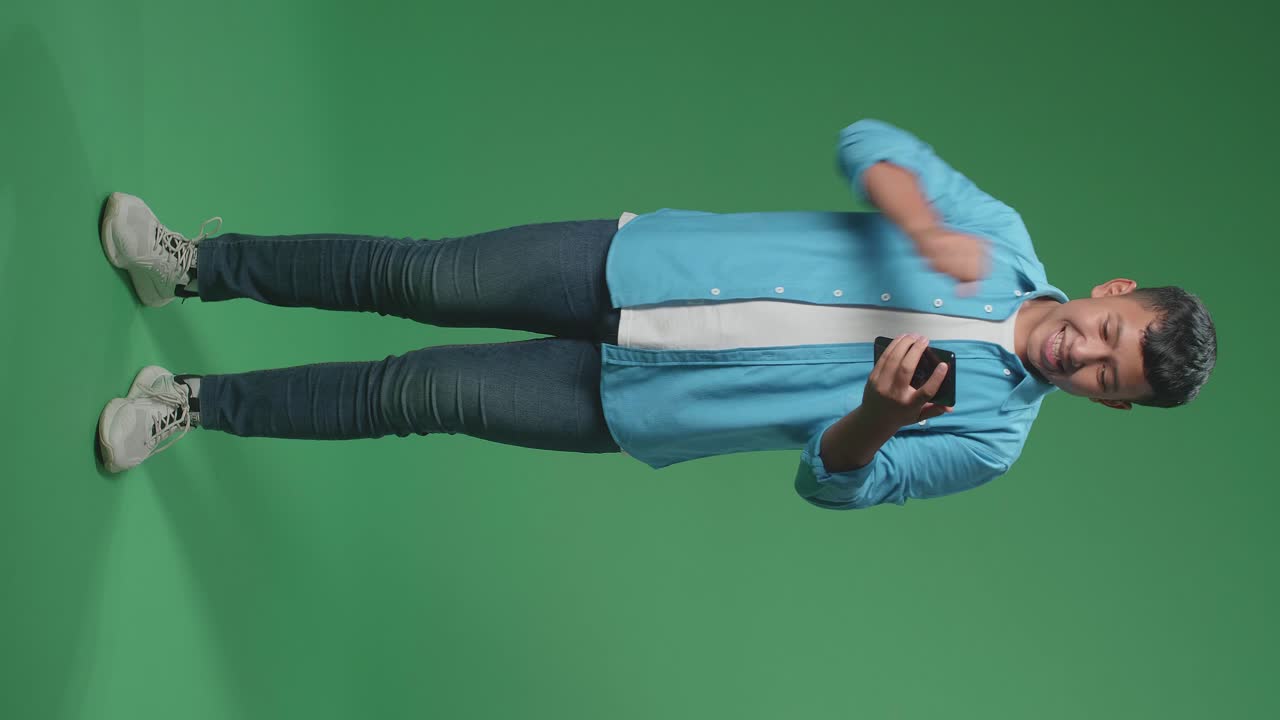 Full Body Of Young Asian Boy Celebrating With Mobile Phone While Standing On Green Screen In The Studio