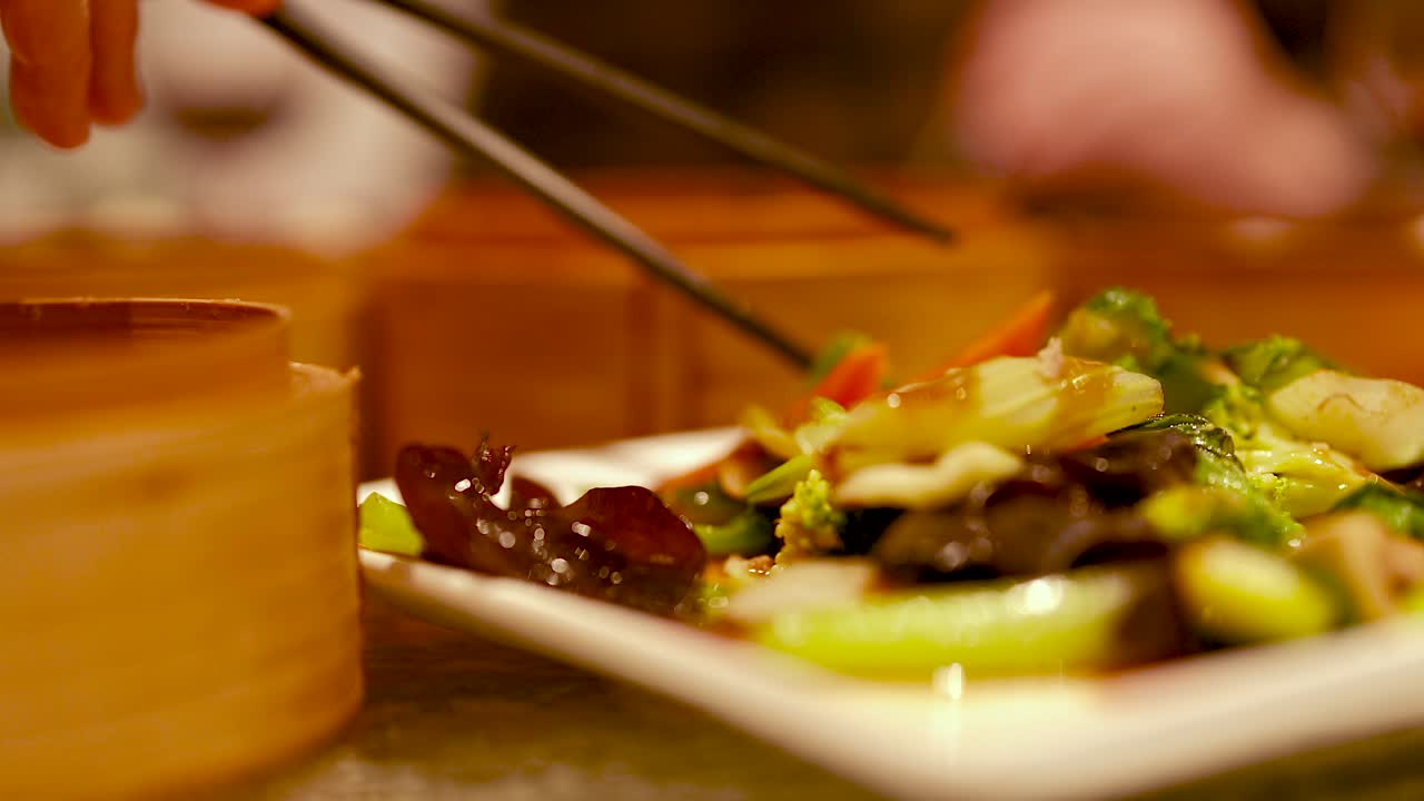 Chopsticks pick colorful stir-fried vegetables from a plate. Warm lighting creates a cozy dining atmosphere