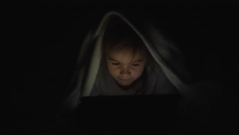 child reading on tablet under a blanket at night