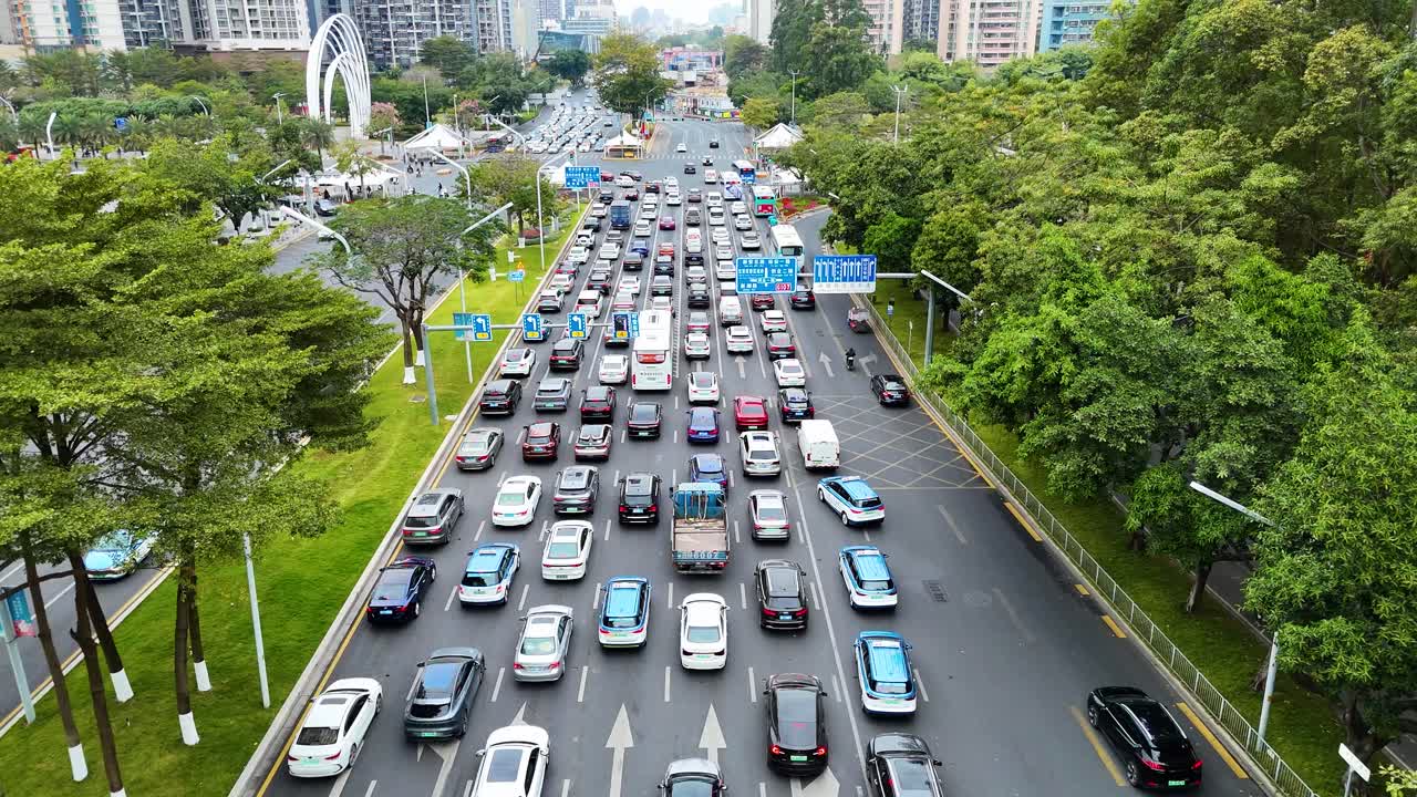 A drone shot capturing the busy urban traffic on a wide road in Shenzhen, China. The video transitions to reveal towering high-rise buildings surrounded by lush greenery.