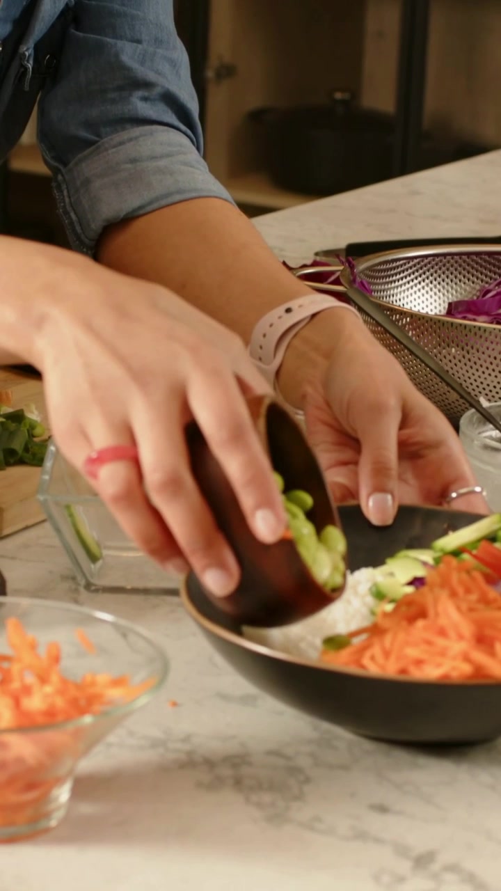 Vertikales Video: Sich vorbeugende Frau im Jeanshemd gießt Edamame in dunkle Schüssel, bereitet Bowl-Mahlzeit zu
