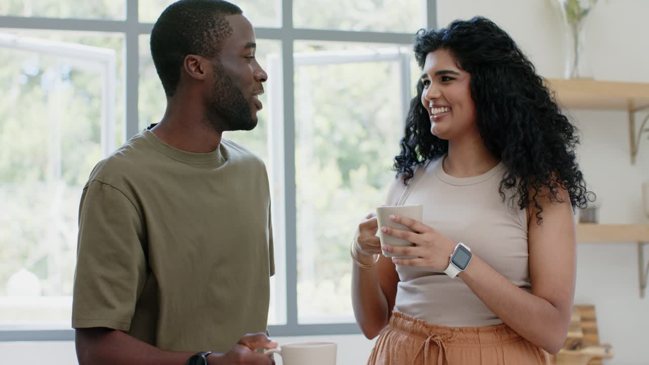 Diverse couple sharing warm moment, meeting eyes and toasting with white mugs in kitchen, embracing