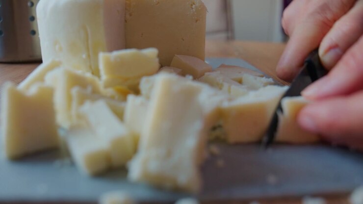 Hand slices hard cheese into cubes on wooden cutting board. Knife glides smoothly through firm texture. Close-up highlights crisp edges and creamy color. Perfect for recipes, food blogs