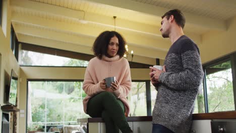 happy diverse couple talking and drinking coffee in kitchen, woman sitting on counter