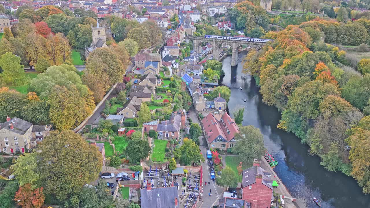 Drone footage pulls away to reveal riverside cottages and terraced gardens, mature trees in autumn foliage, the River Nidd and stone railway viaduct, with historic Knaresborough townscape