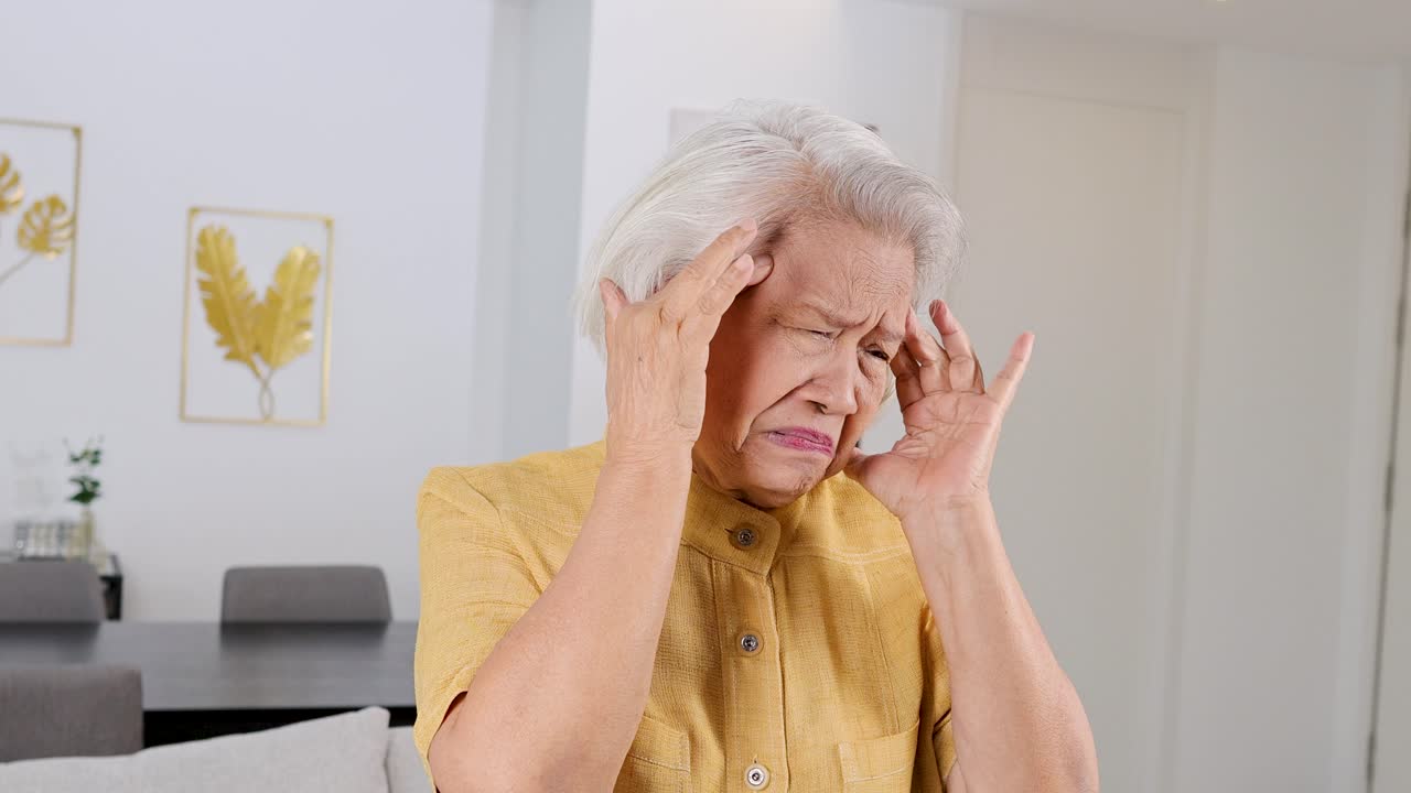 Senior woman in yellow shirt holds head in pain under bright indoor lighting, neutral mood