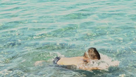 Caucasian-boy-enjoys-swimming-at-sea-of-Kalamata,-Greece,-wearing-a-sea-mask-and-diving-at-clear-waters,-summer-of-2020-,-slow-motion-,-medium-shot