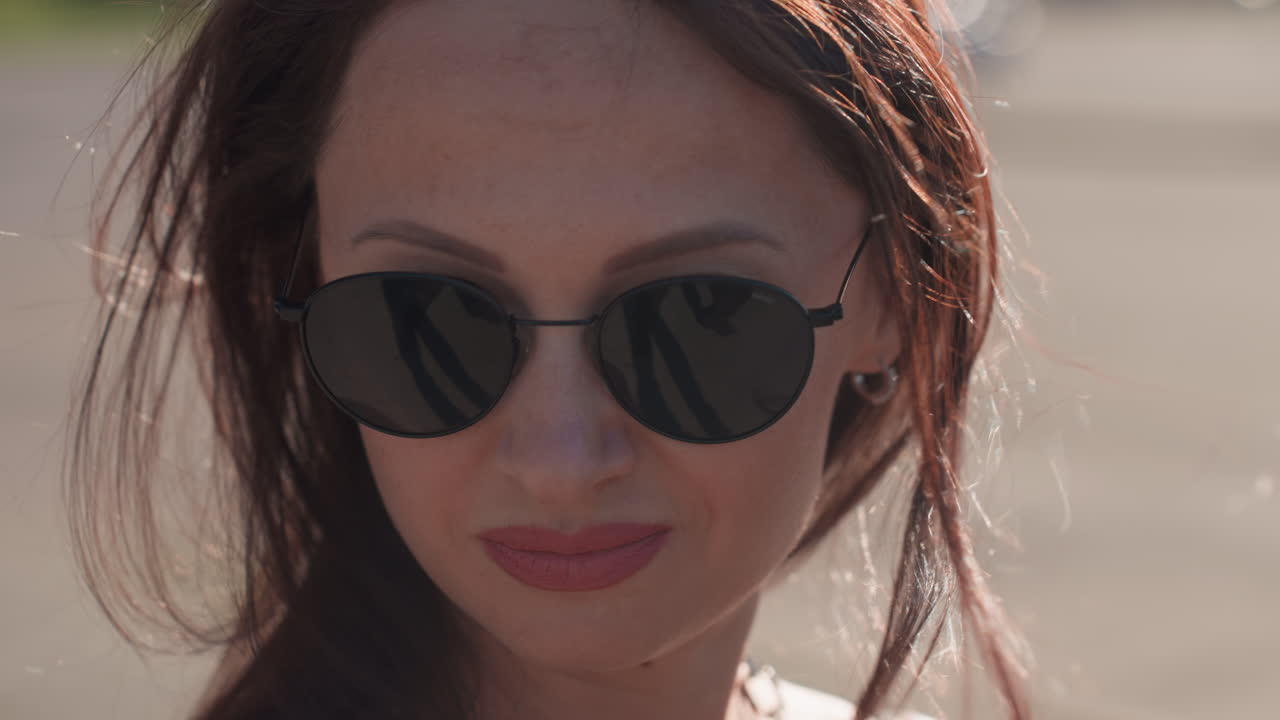 Close up of young woman adjusting sunglasses with hand tattoo visible, hair blowing in wind, outdoor daylight setting showing elegance, personal style and confident feminine expression