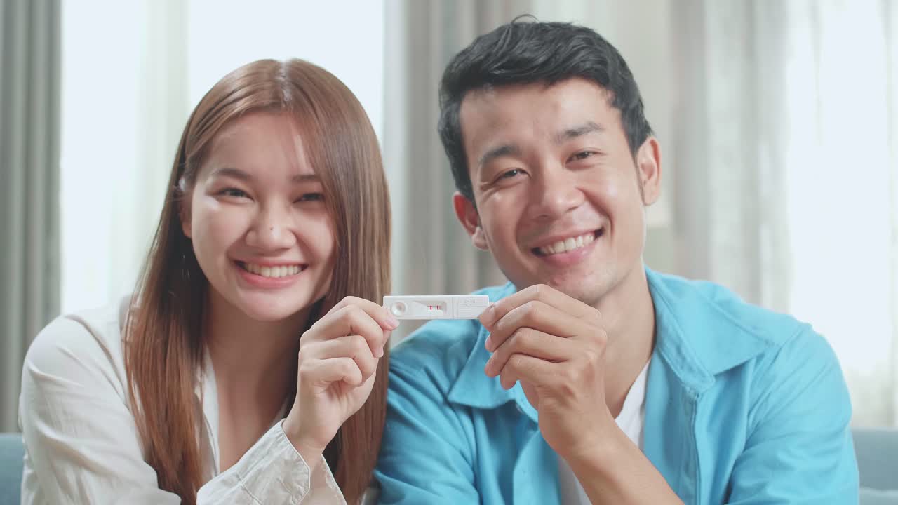 Close Up Happy Asian Couple Holding Positive Pregnancy Test And Smiling To Camera In Living Room At Home
