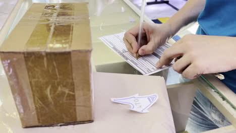 Shot-of-a-woman's-hands-as-she-fills-out-a-form-next-to-a-box-at-the-post-office