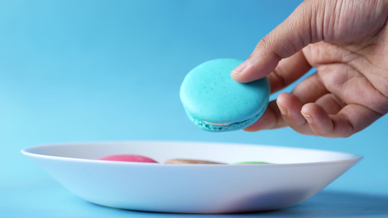 mano sosteniendo un macarón azul azul, con otros macarones de colores en un plato blanco