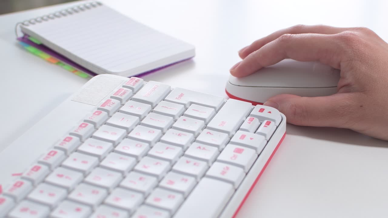 Woman's hand using mouse next to a white keyboard, notepad on white desk.