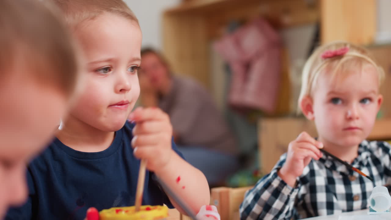 Primo piano di un ragazzino che mescola un pennello in un contenitore di vernice mentre una bambina con una camicia a quadri si concentra sul dipingere accanto a lui, entrambi impegnati in un'attività artistica colorata all'interno di un ambiente creativo in classe