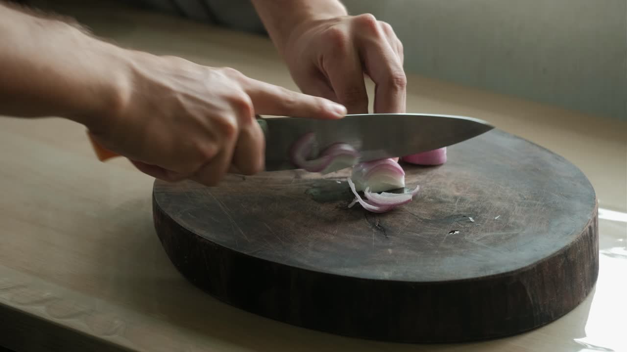 Static shot of hands and chopping knife thinly slicing a red onion on a wooden cutting board.