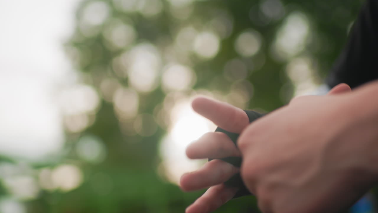 mani all'aperto che indossano un guanto senza dita, primo piano del polso mentre applica una fascia compressiva per la preparazione all'addestramento, sfondo bokeh leggero, retroilluminazione calda, dita tattili che regolano il passante per il pollice, movimento delicato