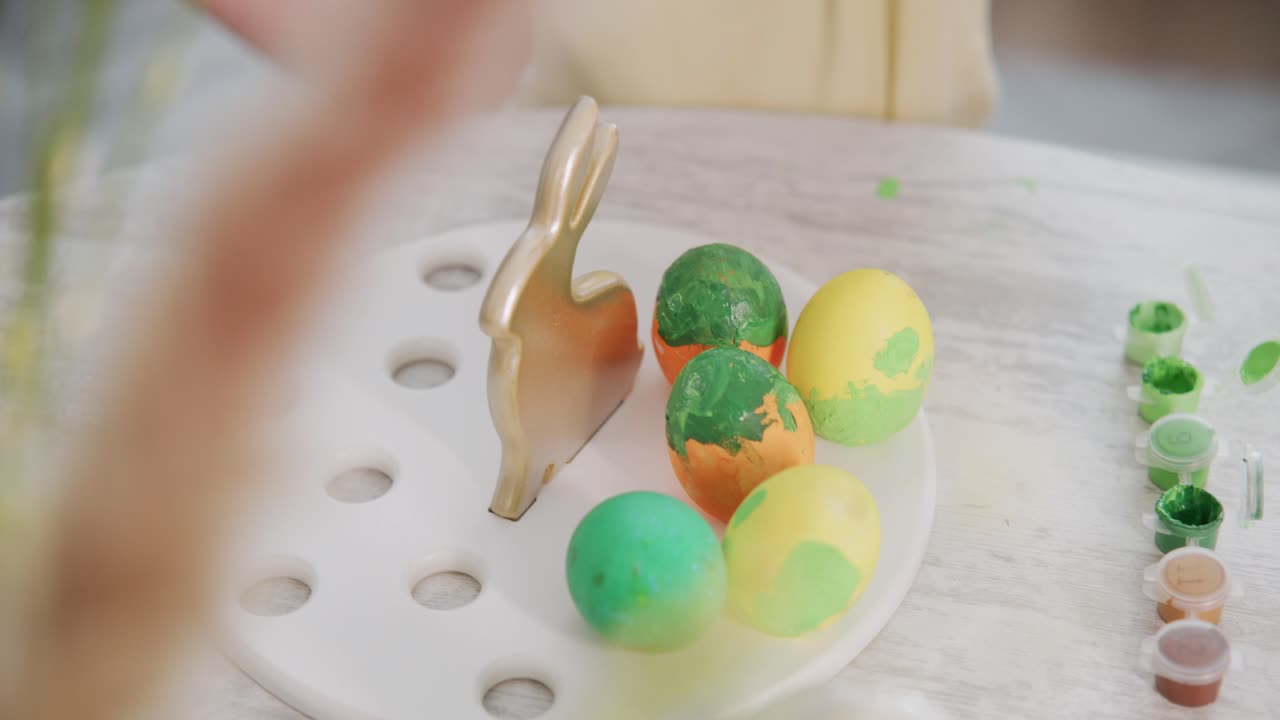 Child paints Easter eggs with vibrant colors. Fingers carefully dip eggs into green and yellow paint. Wooden bunny holds eggs on white tray. Soft focus captures joyful, creative moment