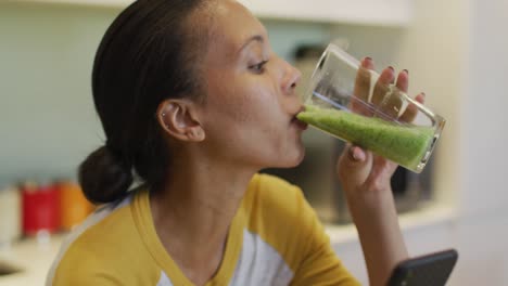 Mixed-race-woman-drinking-healthy-drink-and-using-smartphone-in-kitchen