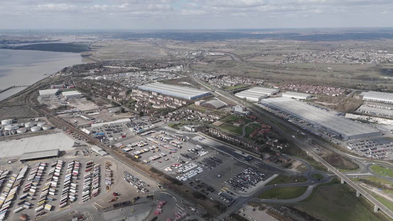 Aerial panorama of vast logistics hub of warehouses, container yards, service roads and rail lines sprawling beside an estuary, highlighting freight transport and supply-chain scale