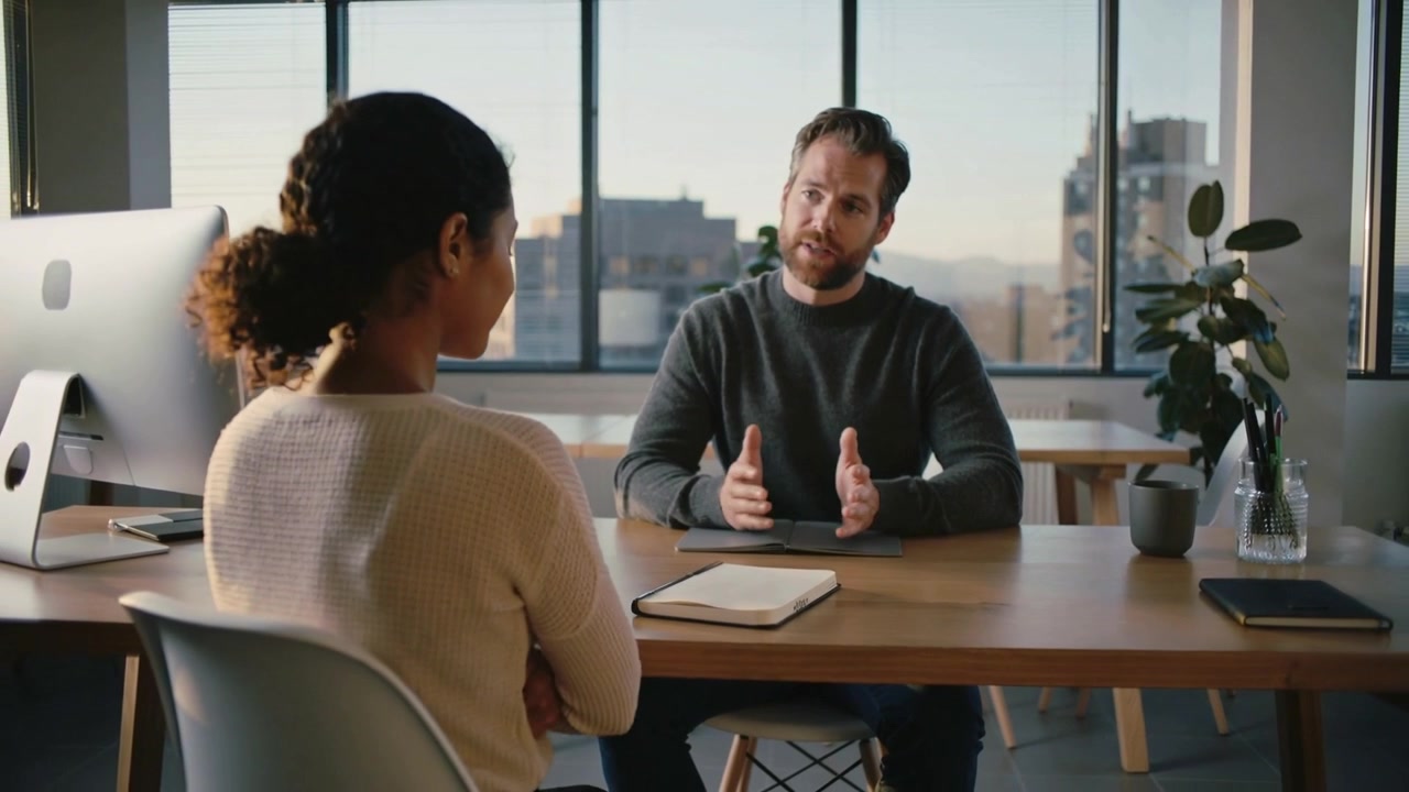 Woman in sweater placing notebook on desk in city office meeting, man in dark sweater explaining