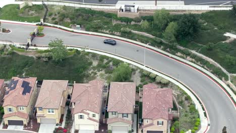 Vista-Aérea-Siguiendo-El-Coche-Eléctrico-Tesla-Negro-Conduciendo-Por-Un-Barrio-Suburbano-Hacia-La-Autopista