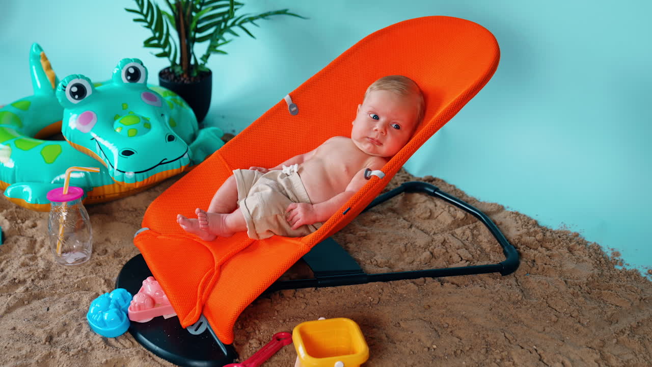 Blond infant boy lies in the orange chair relaxing. Happy vacation on the imitated beach. Side view.