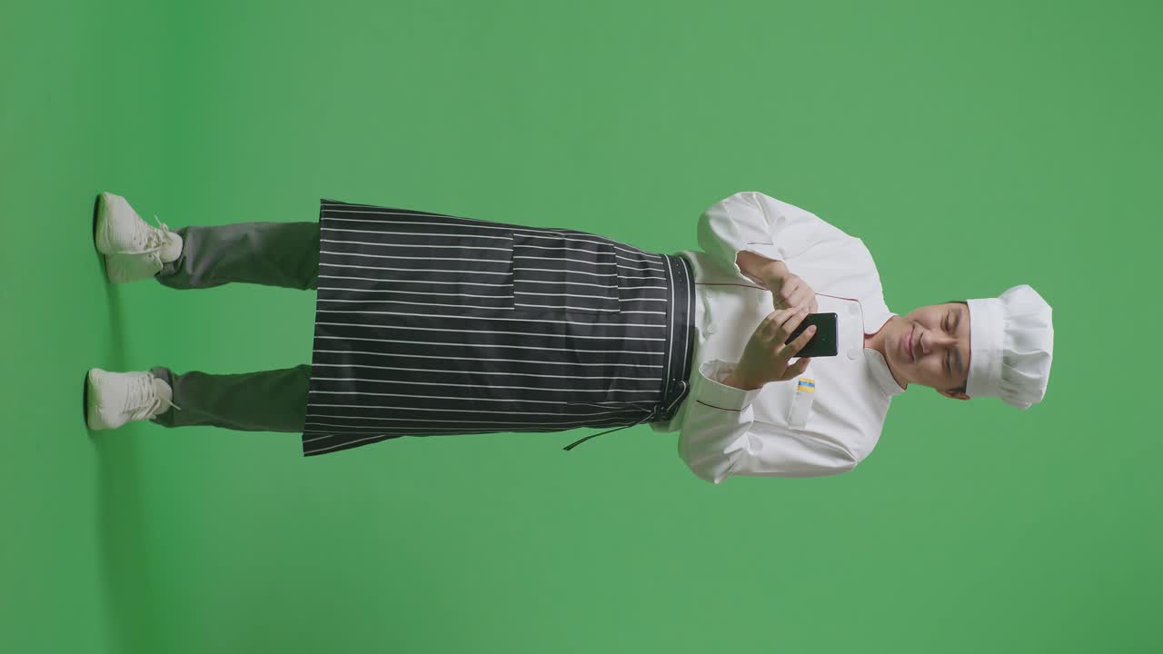 Full Body Of Asian Man Chef Smiling And Using Smartphone While Standing In The Green Screen Background Studio