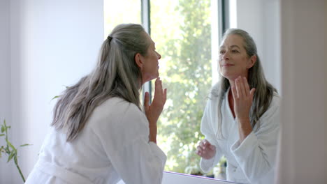 caucasian woman with grey hair applying cream to face, looking in mirror