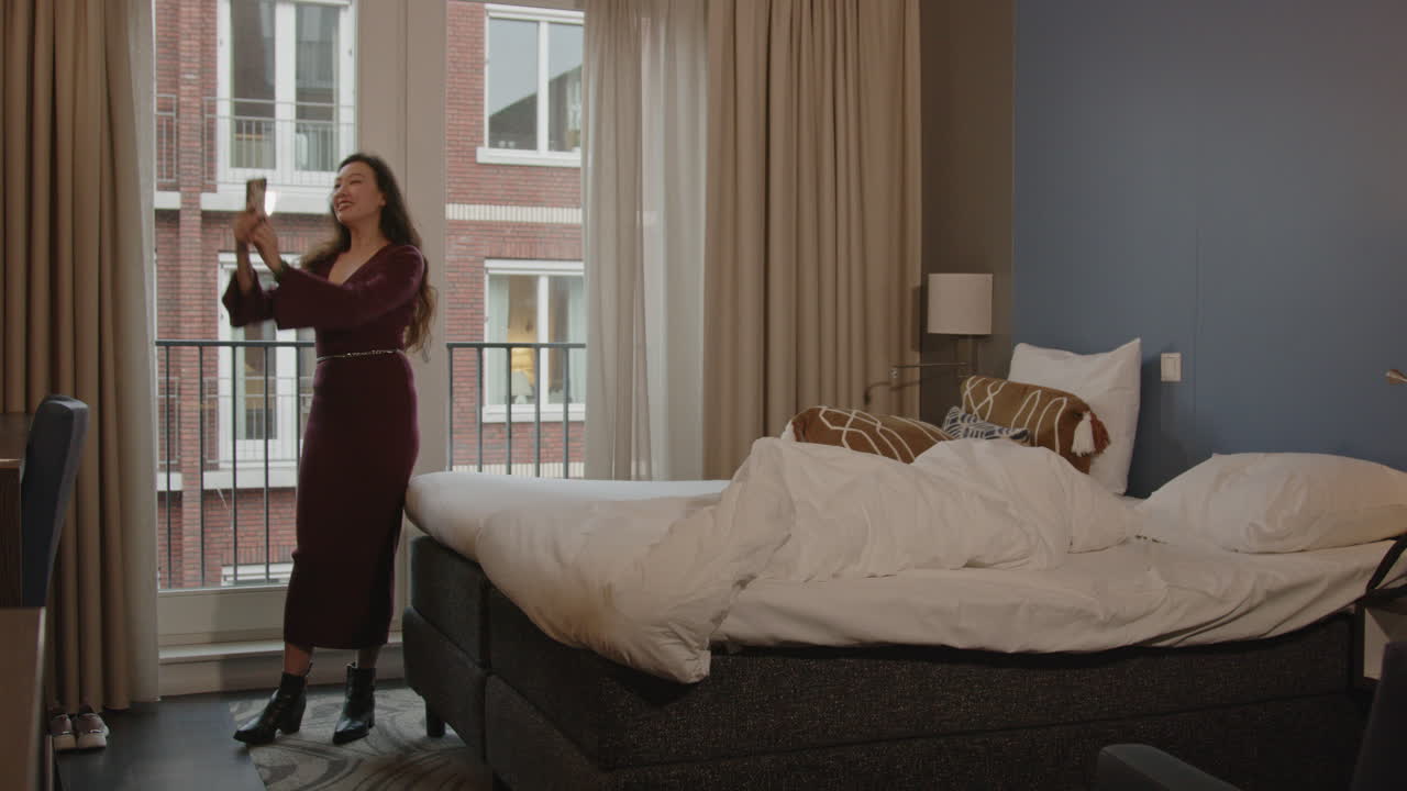 Wide shot of a pretty Asian woman in a hotel bedroom taking a selfie and joyfully walking away