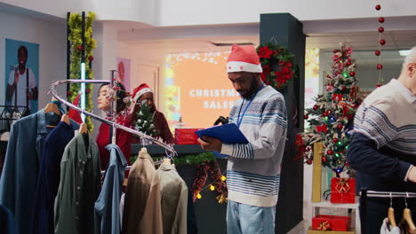 Manager-wearing-Santa-hat-and-holding-clipboard-browsing-through-clothes-racks-at-Christmas-themed-clothing-store,-sorting-out-price-tags-during-holiday-season-promotional-sales