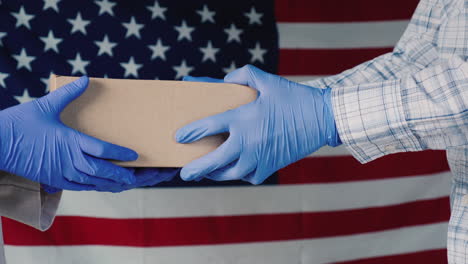 Die-Hände-Eines-Boten-In-Handschuhen-Geben-Das-Paket-Vor-Dem-Hintergrund-Der-Amerikanischen-Flagge-In-Die-Hände-Des-Empfängers