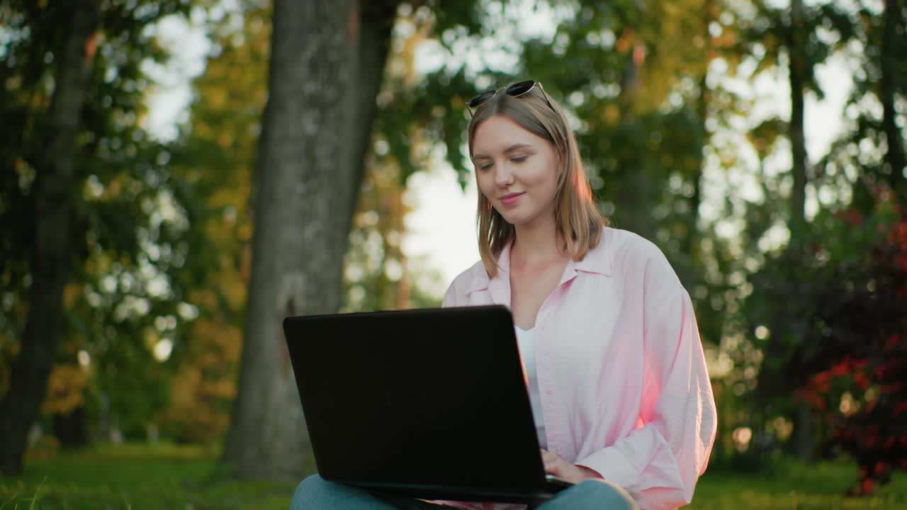 분홍색 을 입은 여자가 잔디에 다리를 어 앉아 노트북을 작업하고, 따뜻한 미소로 사려 깊게 쳐다보고, 자연 빛으로 나무와 초록색을 배경으로합니다.