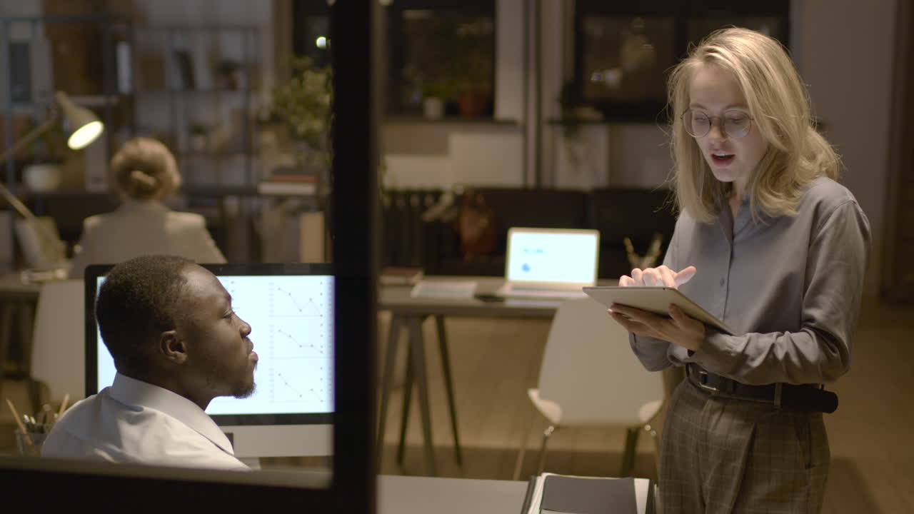 vista trasera del hombre americano sentado en el escritorio mientras habla con una compañera de trabajo que sostiene y mira una tableta en la oficina