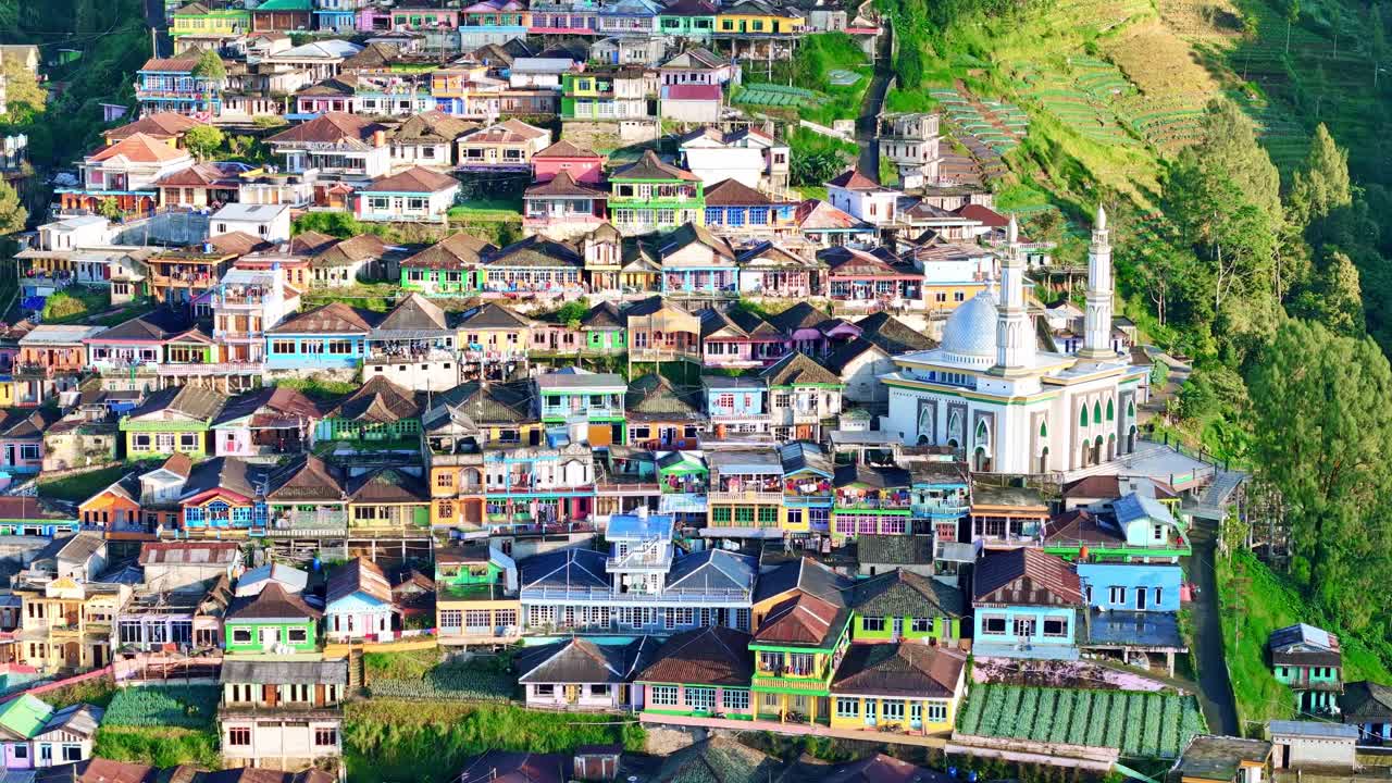 Drone footage of a traditional Indonesian village featuring colorful rooftops, a mosque with twin minarets, and terraced farmlands on the hillside. Nepal Van Java