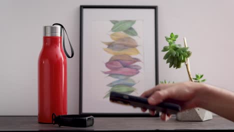 sujeto tomando su teléfono celular, auriculares, reloj inteligente y botella de agua de una mesa decorada con acuarela y planta