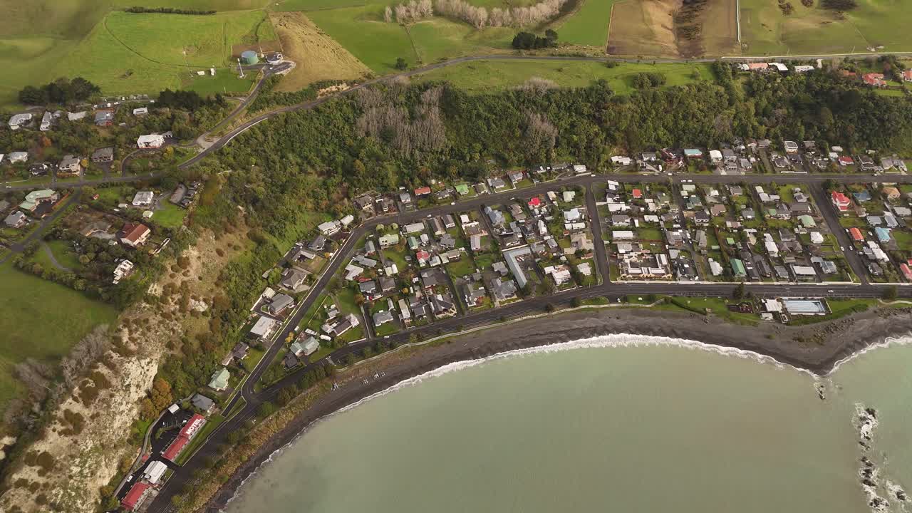 Kaikoura, coastal town on bay with dark sand beach, residential homes between ocean and green hills, South Island, New Zealand. Aerial top-down view
