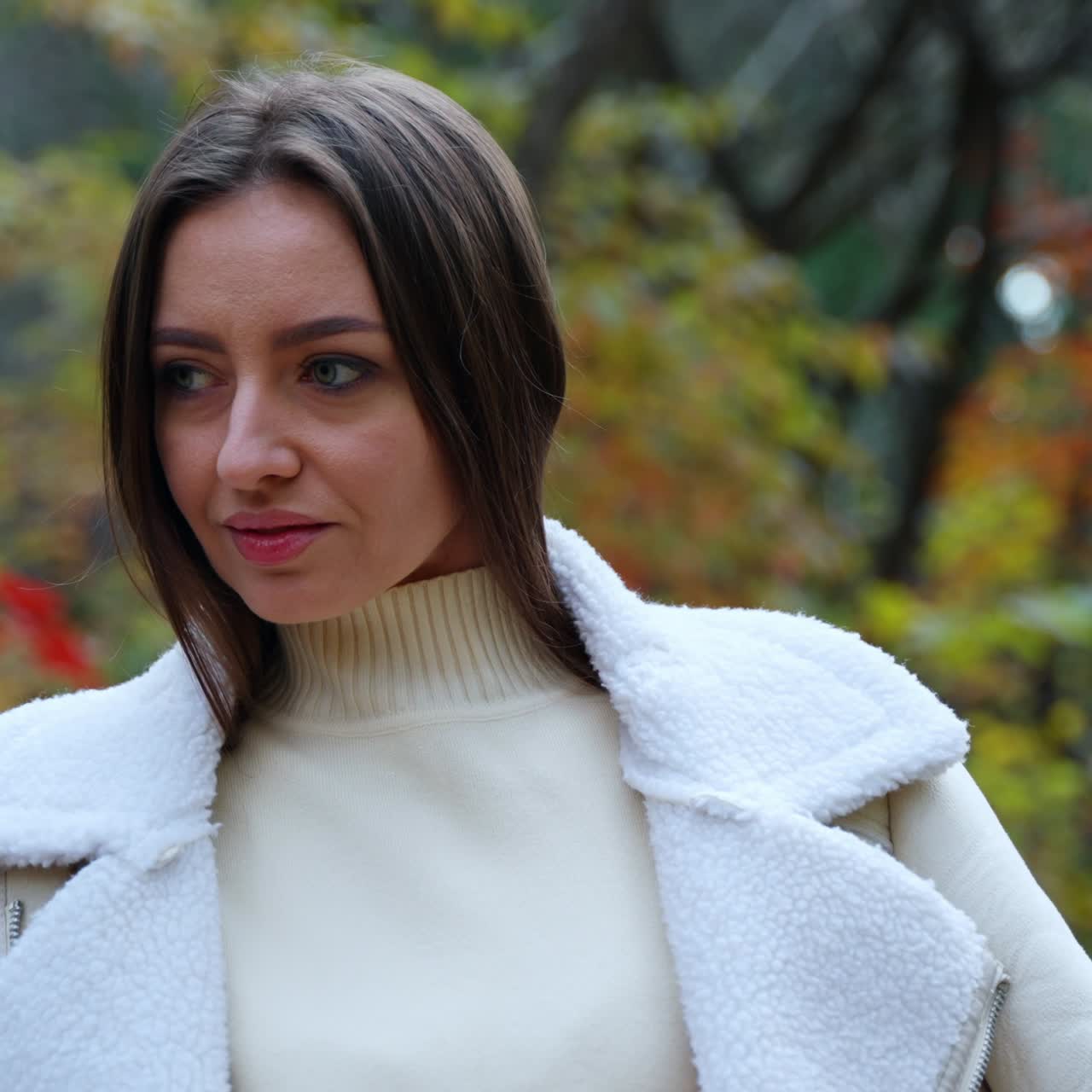 Dark-haired Caucasian woman stands outdoors with jacket on her shoulders and hair hidden under it. Lady posing in the beautiful autumn park