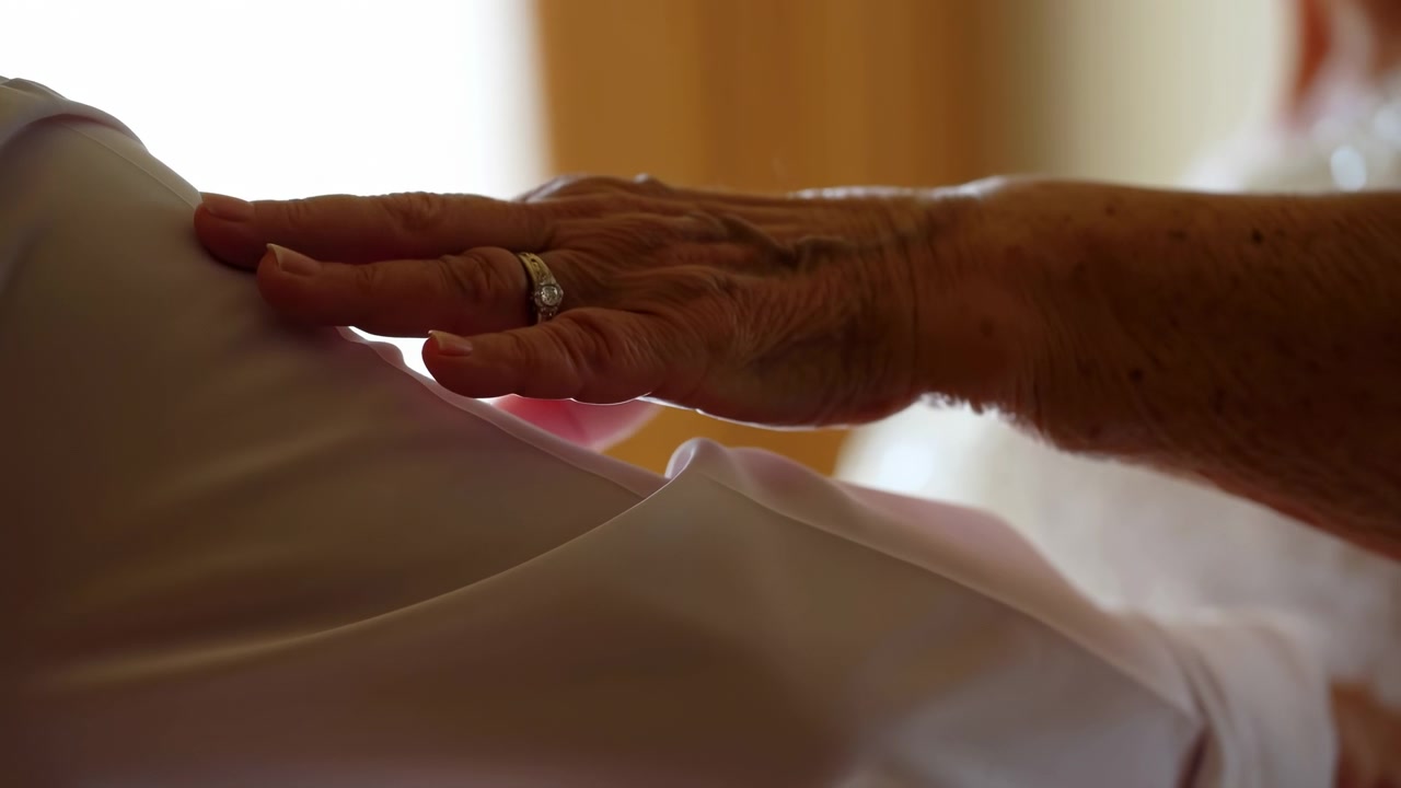 Close-up view shows a ringed hand resting on and slowly moving over a clothed shoulder, with soft daylight, warm interior tones, shallow focus, and gentle repeated contact.