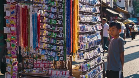young boy stands observing a colorful display of religious souvenirs at a bustling street market stall, captivated by the variety of items