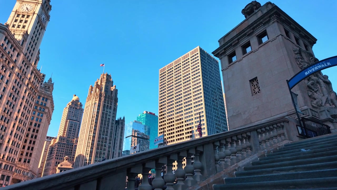 Torri gotiche e insegna della passeggiata lungo il fiume da una scalinata in pietra, centro di Chicago. Angolazione bassa di una balaustra ornata, torre dell'orologio e skyline di grattacieli sotto una luce dorata.