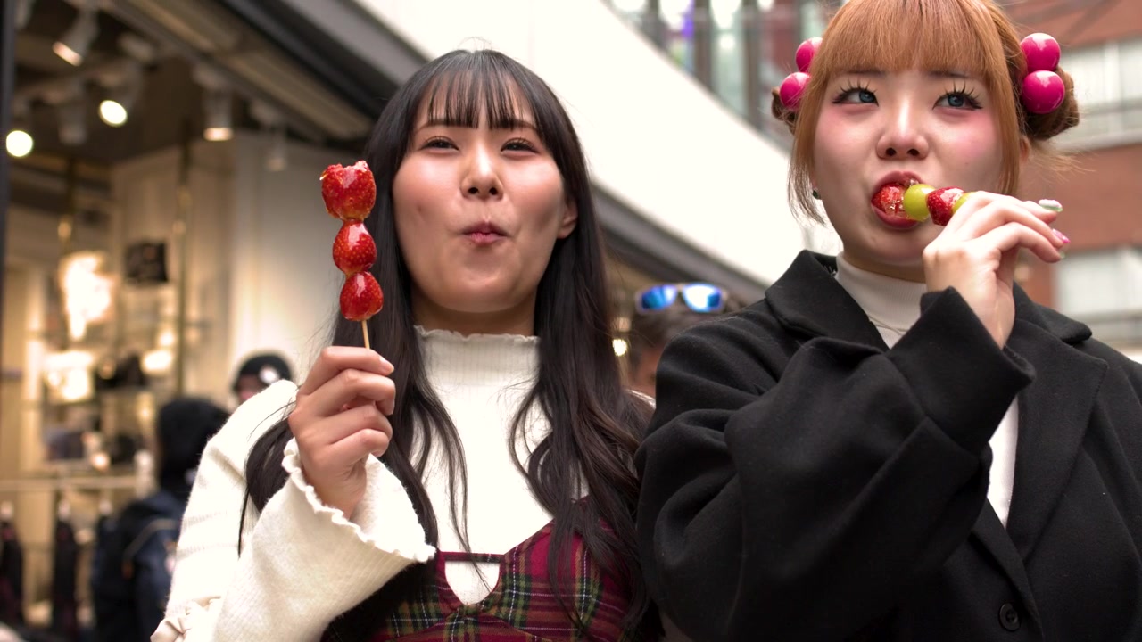 Due amiche alla moda mangiano spiedini di frutta candita mentre passeggiano per un affollato quartiere commerciale di Tokyo.