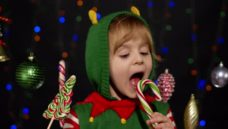 Child-kid-girl-in-Christmas-elf-Santa-Claus-helper-costume-licking-candy-lollipop-caramel-sweets