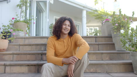 Retrato-De-Un-Hombre-Birracial-Feliz-Con-El-Pelo-Largo-En-Los-Escalones-De-Un-Jardín-Soleado,-Cámara-Lenta,-Espacio-Para-Copiar