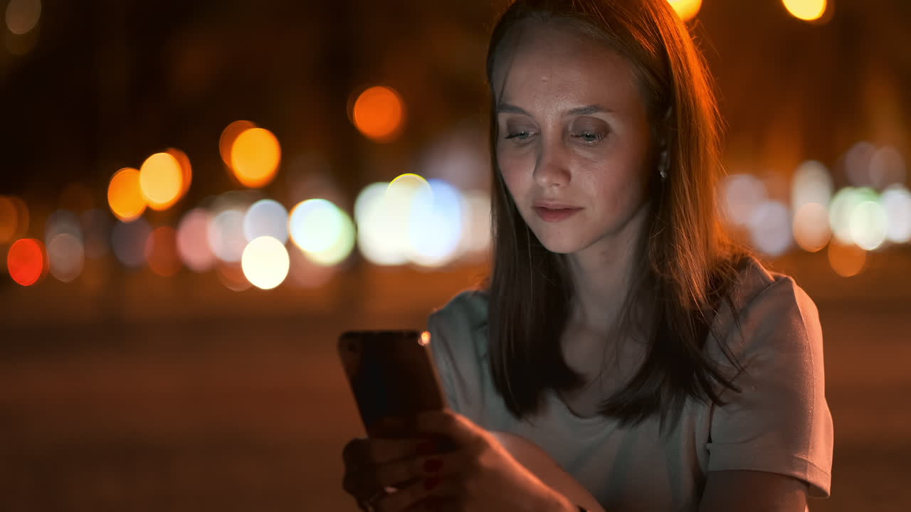 At night a young girl sits on the grass in a big city and looks into the smartphone screen writes a message and maintains her personal blog.