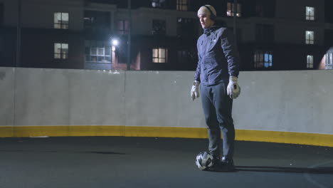 man in headphones wearing joggers skillfully traps incoming ball on outdoor court at night with urban residential buildings and bright lights in the background