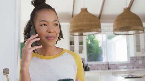 African-american-woman-holding-coffee-cup-talking-on-smartphone-at-home