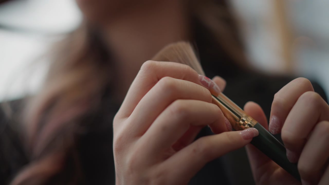 Tight low-angle close-up shows hands gripping makeup brush, adjusting hold, tapping handle, and fluffing bristles in preparation for cosmetic application with soft warm background blur