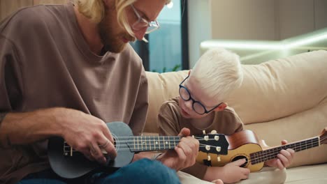 un niño albino con cabello blanco y gafas azules mira a su padre tocar la guitarra y el ukulele y trata de repetir después de él mientras está sentado en el sofá de un apartamento moderno. niño albino aprende a tocar un instrumento musical con su padre en casa